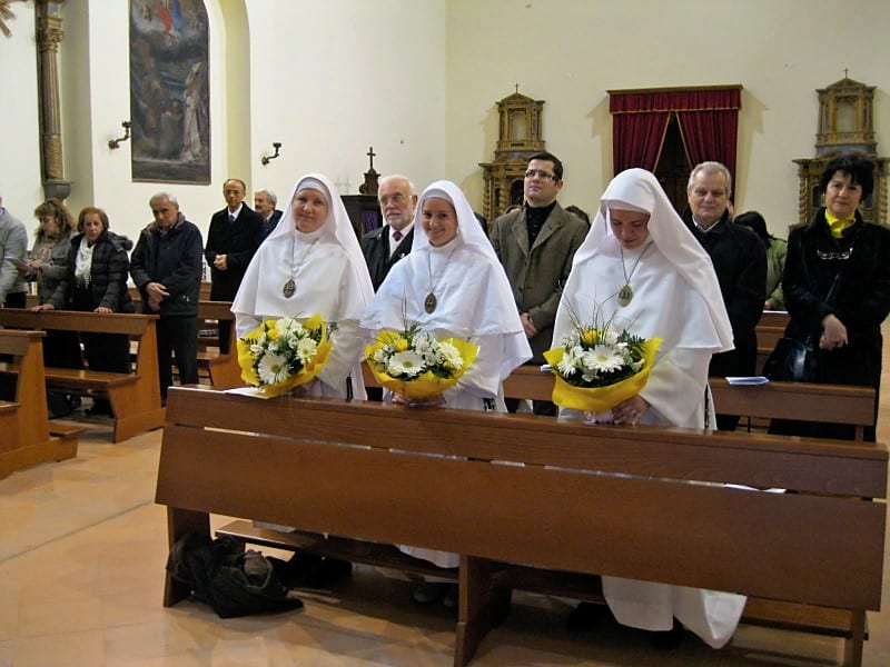 Four sisters in white habits holding yellow and white flower bouquets standing in a row inside a church before the ceremony.