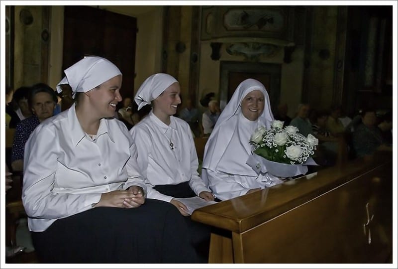 Three sisters seated in a church pew, smiling, two in white habits and one in an older dark habit holding white roses.