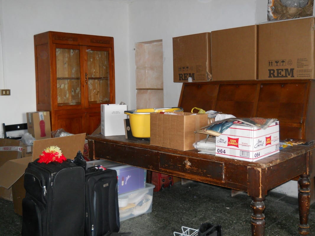 A large room on moving day with a long wooden table buried under cardboard boxes, suitcases, and other belongings.