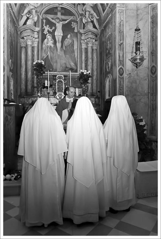 Three sisters in white habits kneeling with their backs to the camera before a frescoed altar in a dimly lit church.