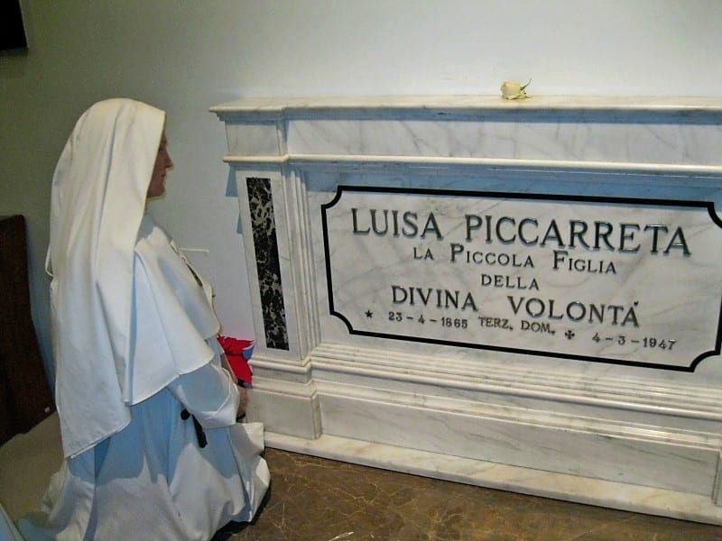 A sister in a blue-and-white habit kneeling at the tomb of the Servant of God Luisa Piccarreta, her name and dates inscribed on the marble.