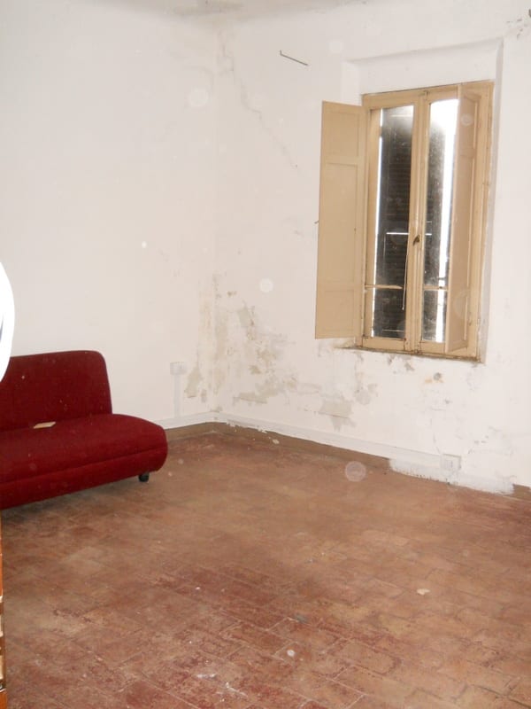 An empty room with peeling walls, bare floorboards, and a single red sofa beneath an open shutter window.