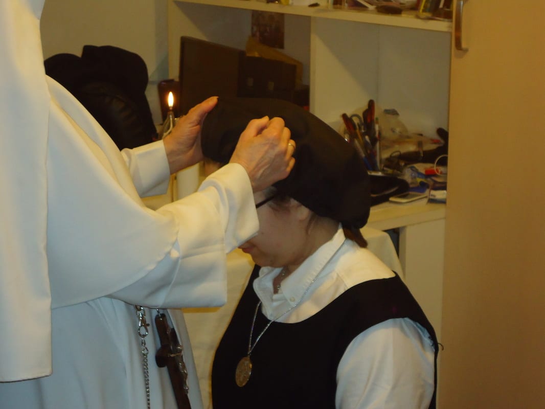 A black veil being placed on the head of a sister in a dark habit by another sister's hands, in a small room with a lit candle.