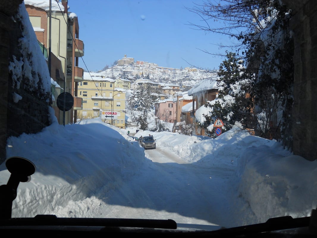 Snow-covered rooftops and a street scene viewed through a windshield, with a rocky hillside in the background.