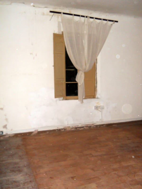 An empty room with white walls, bare floorboards, and a single window with thin curtains.