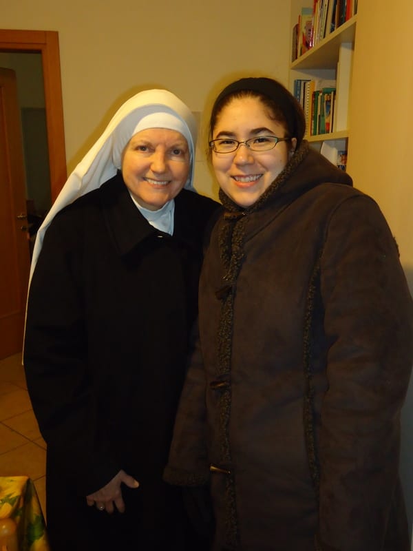 A sister in a white habit and a young woman in a brown coat standing together and smiling indoors.