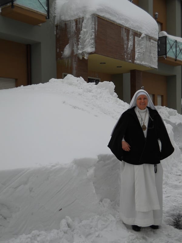 A sister in a black habit and cape standing before a massive snow drift piled against a building exterior.