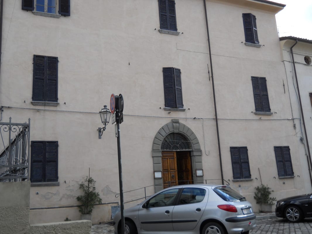 Exterior of a large pale stone building with dark shuttered windows and a central arched doorway, a car parked in front.