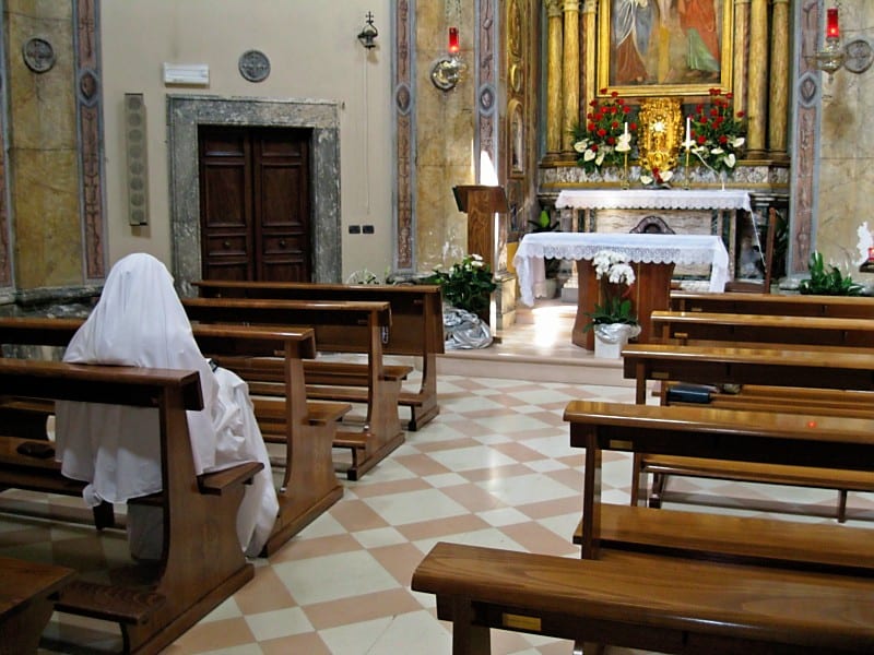A sister in a white habit seated alone in a nearly empty church, facing the decorated sanctuary during adoration.