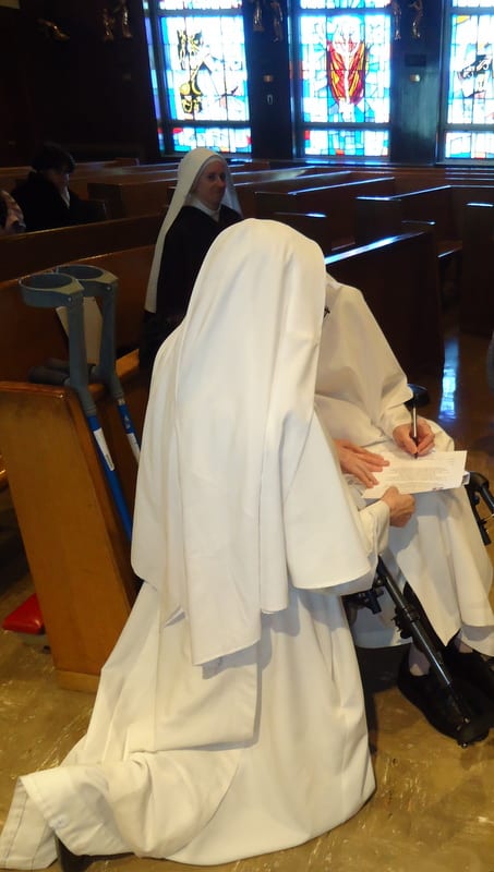 A sister in a white habit kneeling and writing in a book, her head bowed, in a church pew.