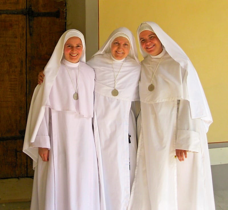 Three sisters in white habits standing together smiling outdoors, arms around one another.
