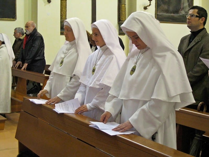 Three sisters in white habits standing at a pew reading from papers held in their hands, with lay attendees behind them.