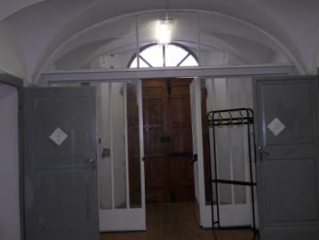 A vaulted corridor or entrance hall with grey double doors, a wooden inner door open to light beyond, and a metal rack against the right wall.
