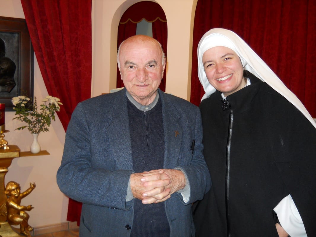 An elderly man in a dark jacket and a sister in a white habit standing side by side smiling in a room with red drapery.