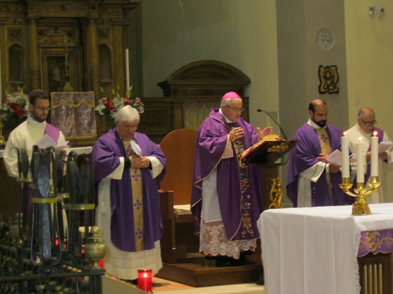 A bishop in purple vestments at the altar with two concelebrants, flower arrangements flanking the sanctuary.