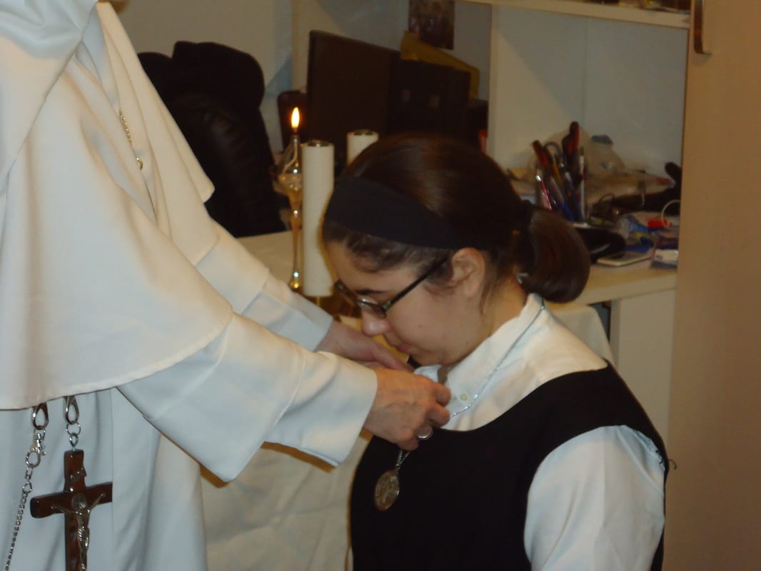 A sister in a dark habit bowing to kiss a medal being placed around her neck by another sister's hands.