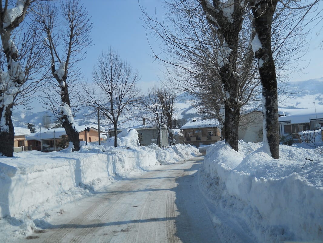A snow-lined road flanked by bare trees stretches through a village blanketed in deep snow, hills visible in the background.