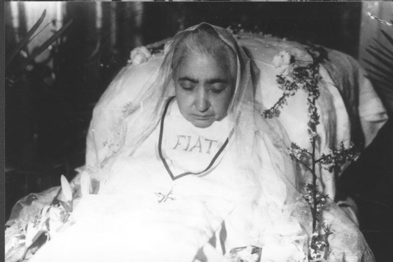 Black-and-white photograph of the Servant of God Luisa Piccarreta in repose, wearing a white veil and the word “FIAT” on her chest, with a rosary.