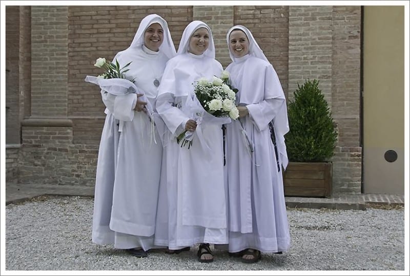 Three sisters in white habits and veils stand outdoors holding bouquets of white flowers, smiling for the camera.