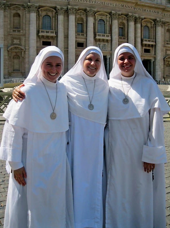 Three sisters in white habits smile together in St. Peter's Square, the facade of St. Peter's Basilica visible behind them.