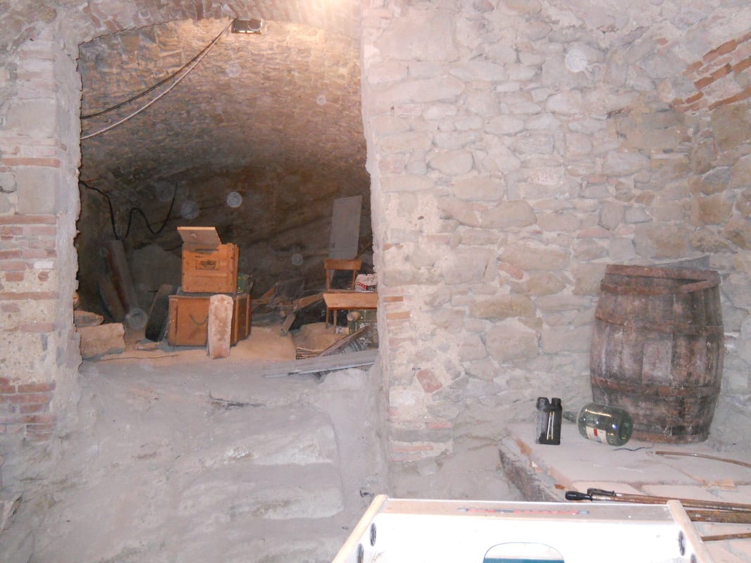 A low stone-vaulted cellar room with rough masonry walls, scattered wooden boxes and an old barrel in the corner.