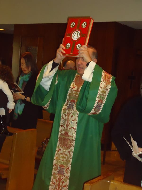 A priest in a green chasuble raises a red-covered book during the Liturgy of the Word at an indoor Mass.