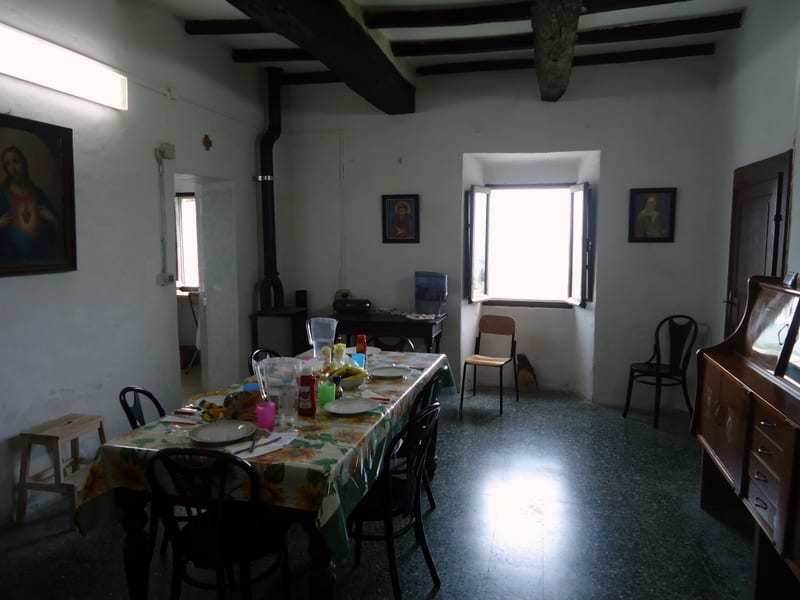 A simply furnished dining room with exposed wooden ceiling beams, a long table set with a colorful cloth, religious images on the walls and a sideboard along one side.