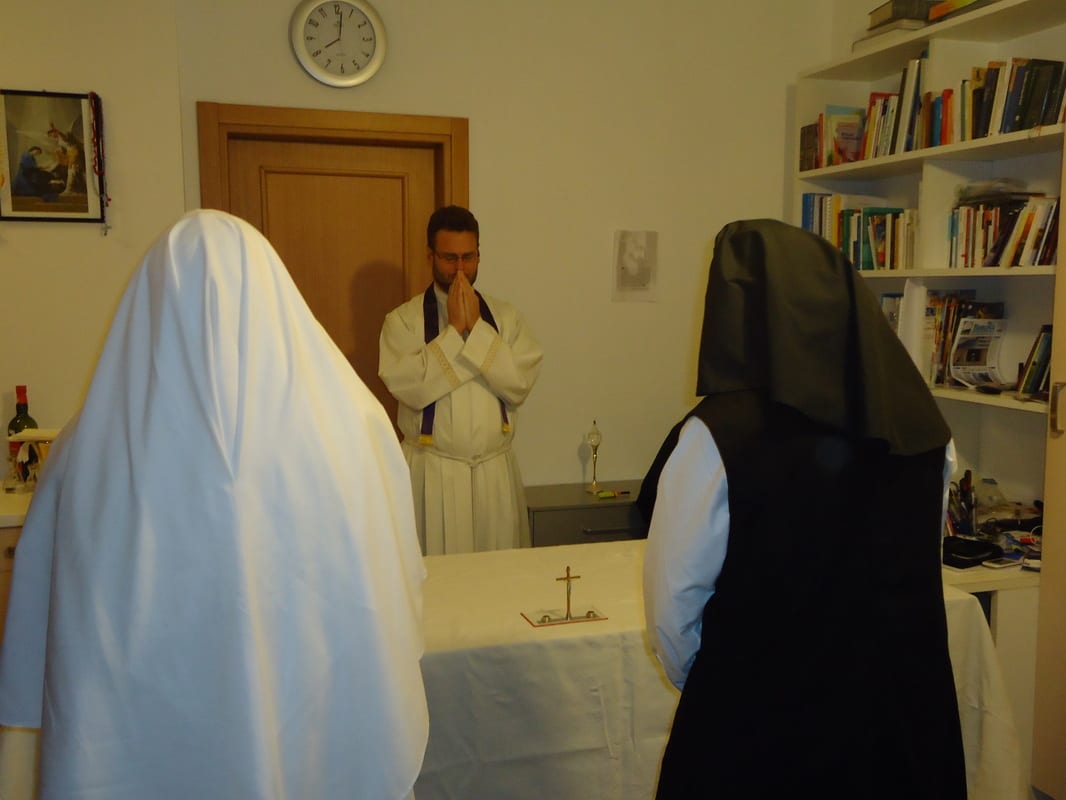 A priest in a white alb stands facing a small altar inside a room, while a sister in a white habit and a sister in a black habit stand before him with heads slightly bowed.
