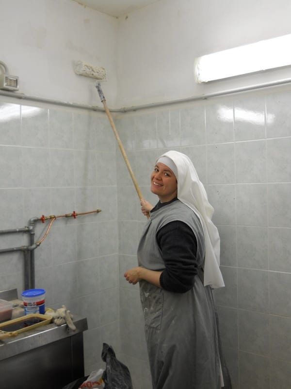 A sister in a grey apron and white veil uses a long-handled brush to scrub a tiled wall in a kitchen or utility room.