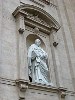 Stone niche statue of a robed male saint, set in a classical architectural surround on a large stone facade.