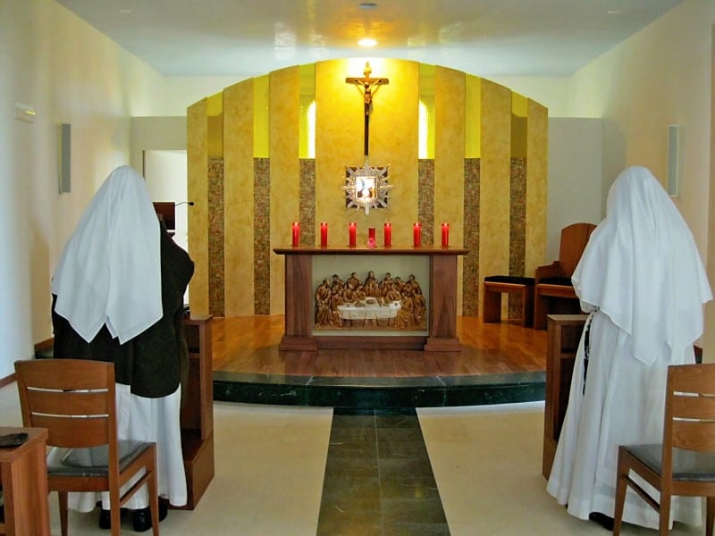 Two sisters kneel in prayer before a tabernacle flanked by red candles and a carved relief panel in a small, bright chapel.