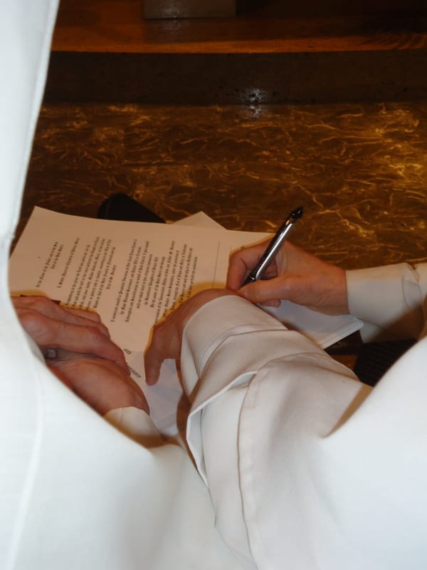 Close-up of a person in a white habit signing a document with a pen, the paper resting on a wooden surface.