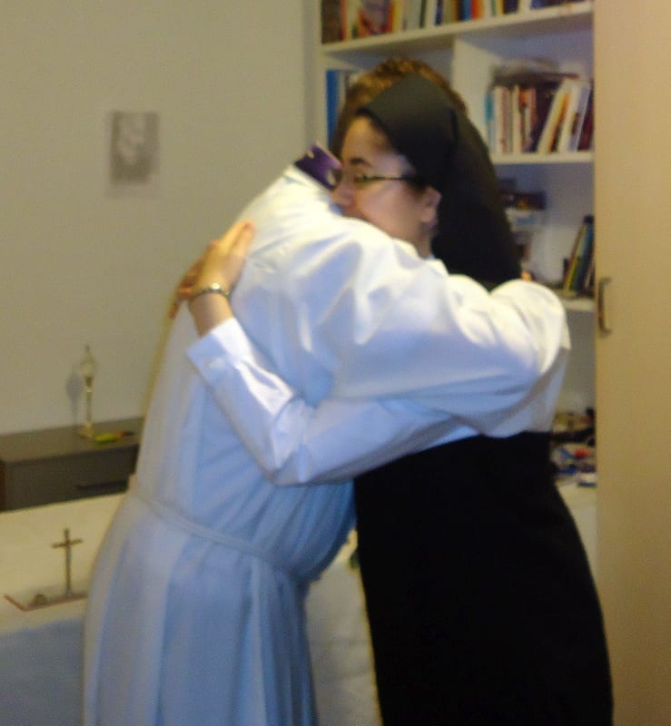 Two people embrace warmly in a small room lined with bookshelves, one wearing a white habit and the other in dark clothing.