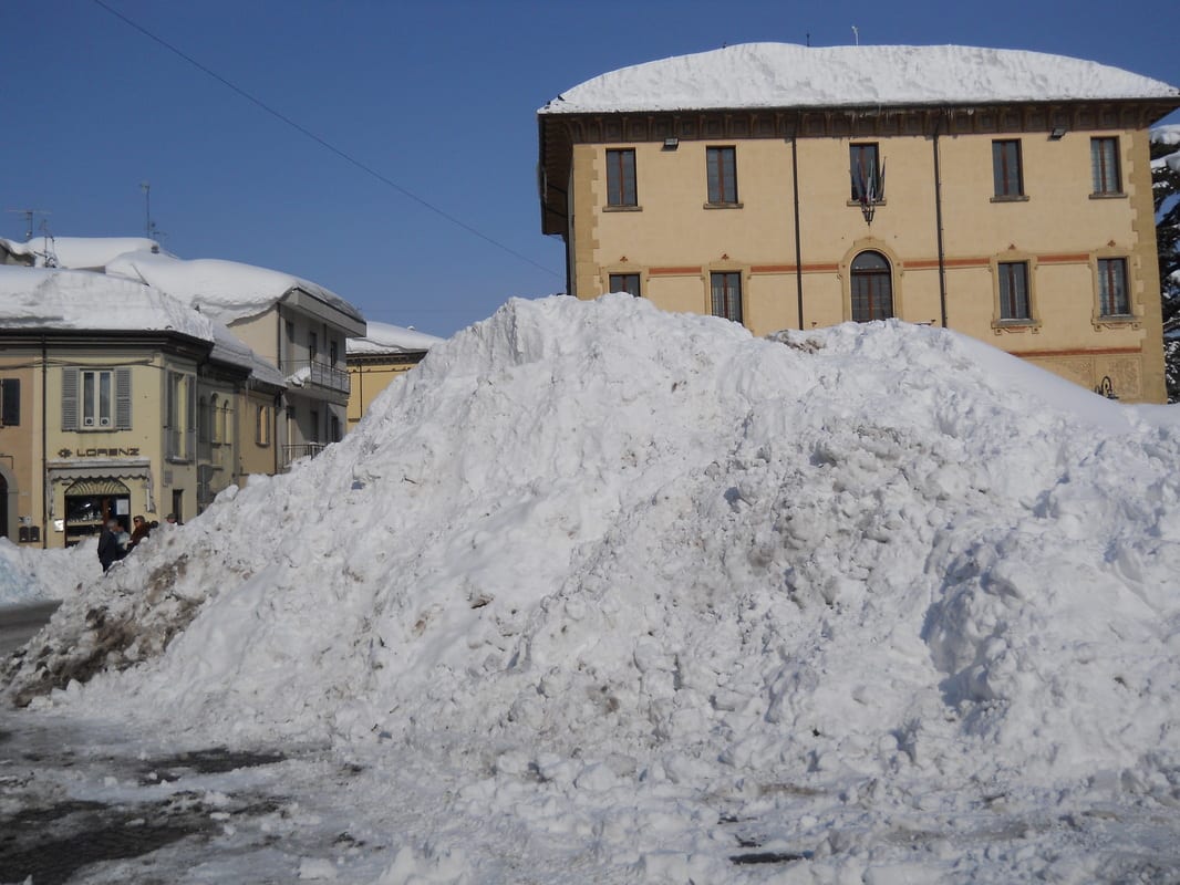A large mound of plowed snow piled in a town square, with pale yellow and ochre buildings visible across the street under a clear blue sky.