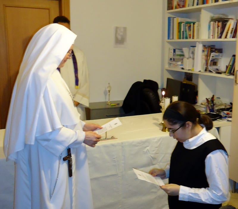 A sister in a white habit holds a piece of paper while a younger woman in dark clothing reads from her own paper, with a bookshelf-lined room behind them.