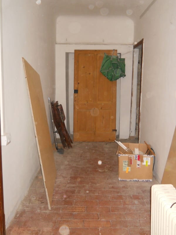 A narrow hallway under renovation with bare brick floor, wooden door at the end, loose boards leaning against the wall and a cardboard box on the floor.