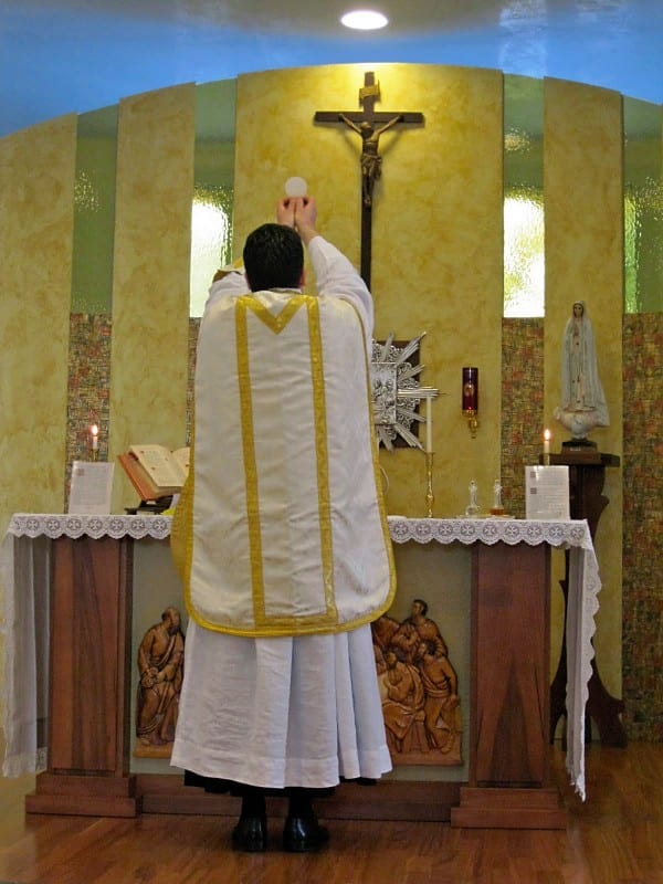 A priest in white vestments stands at an altar with arms raised during Mass, a crucifix and a statue of Our Lady visible behind him.