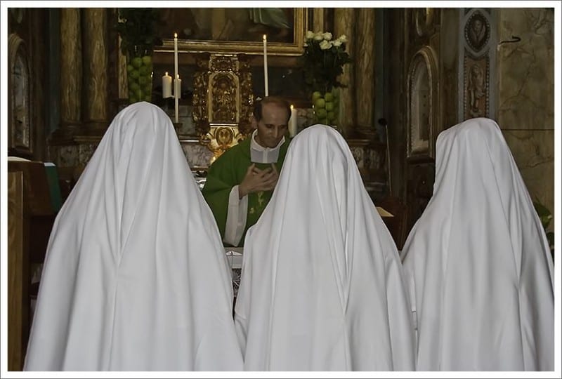 Three sisters in white habits kneel facing the ornate high altar of a historic church while a priest in green vestments celebrates Mass.