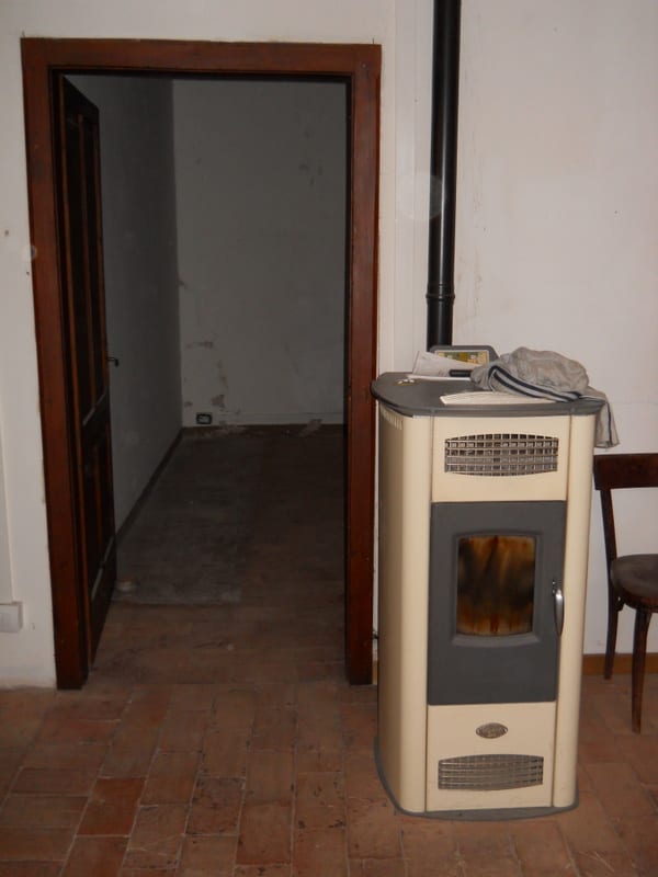A small wood-pellet stove in the corner of a room, with a doorway opening into an adjacent empty room.