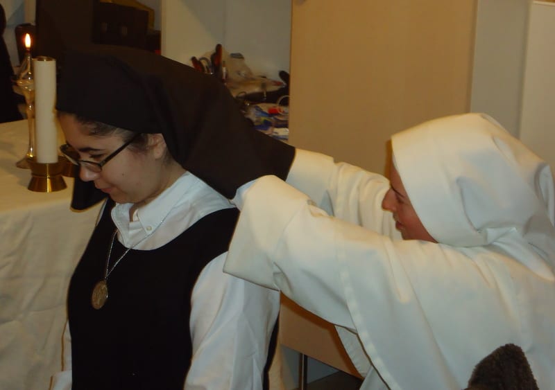 A sister in a black habit adjusts the veil or habit of another sister by candlelight, with an altar table in the background.