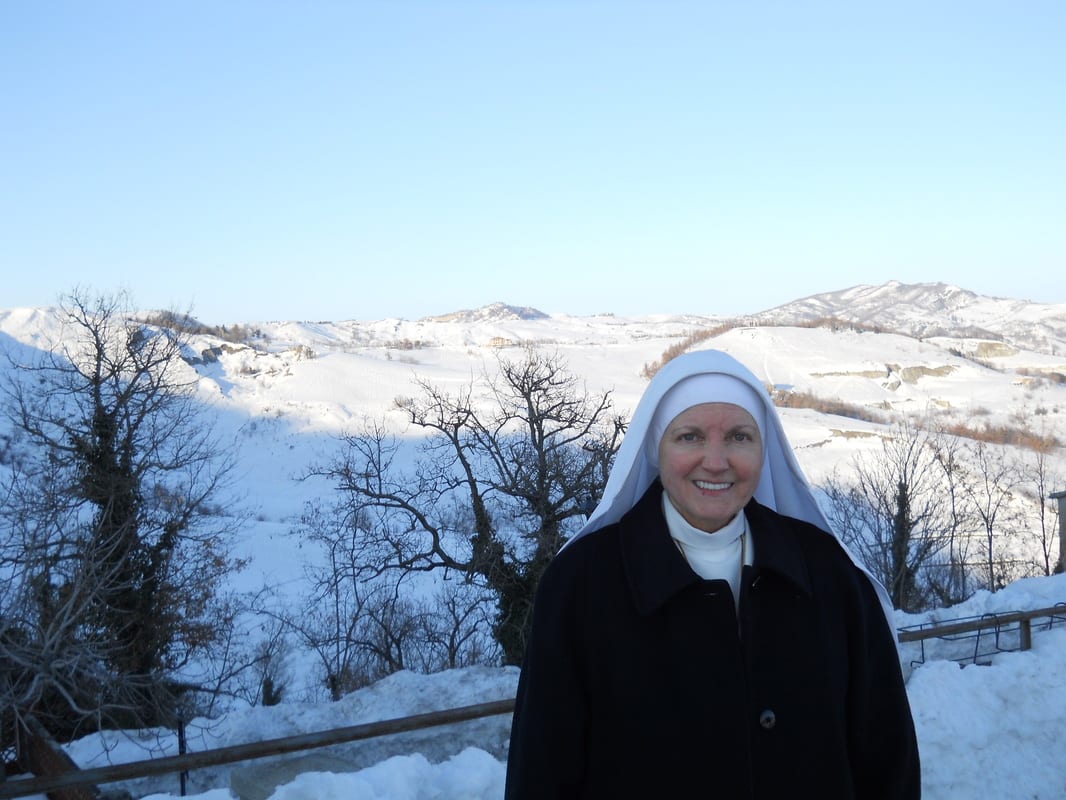 A sister in a black winter coat and white veil stands smiling on a terrace or balcony with a wide snow-covered valley and bare-branched trees stretching behind her.
