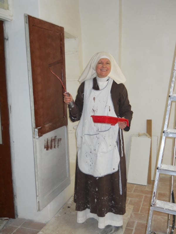 A sister in a brown habit and white apron stands smiling in a partly renovated room, holding a paint roller, with a ladder behind her.