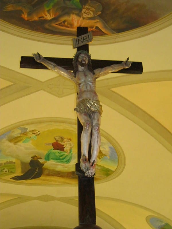 A large crucifix mounted against a painted arch in a church apse, with a devotional mural of saints visible in the lunette behind it.