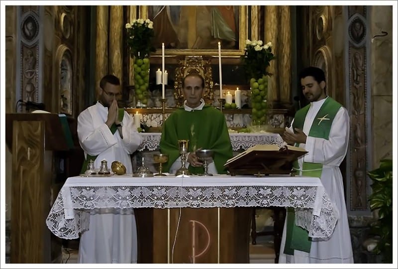 A priest and two servers stand behind an altar set with candles and a missal in an ornate historic church during Mass.