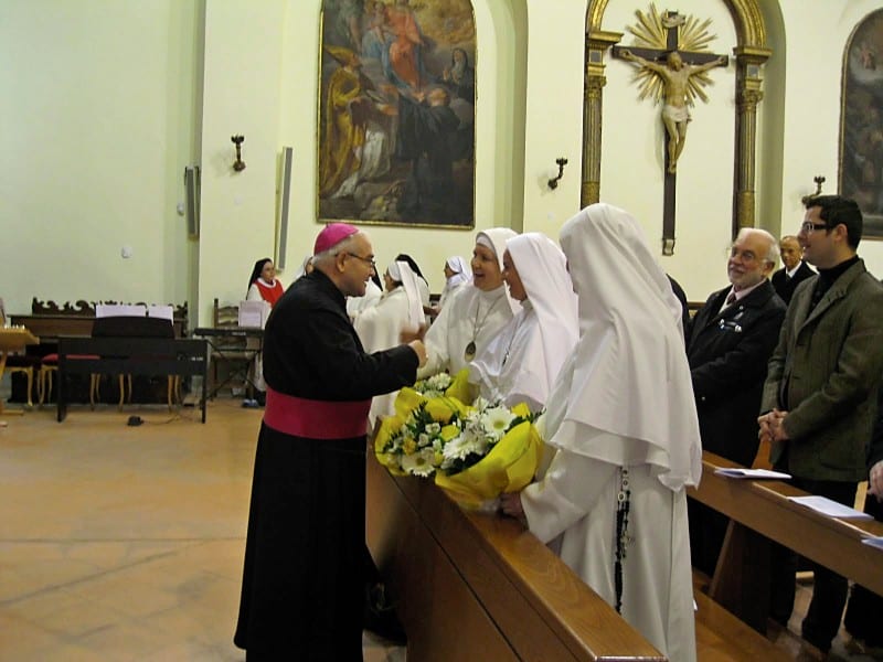 A bishop in purple choir dress and pectoral cross presents a bouquet of flowers to a sister in a white habit inside a church, with other religious and laity looking on.