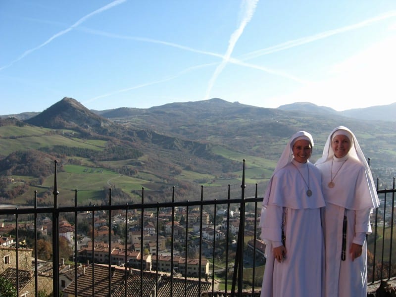 Two sisters in white habits stand on a scenic terrace overlooking a wide valley of green and brown hills under a blue sky.