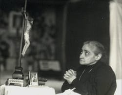 Black-and-white photograph of the Servant of God Luisa Piccarreta seated in profile, hands clasped in prayer before a crucifix and small devotional images.