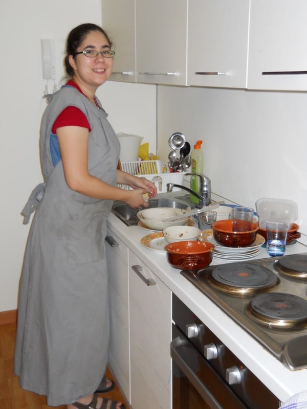 A young woman in a grey apron washes dishes at a kitchen sink, smiling over her shoulder.