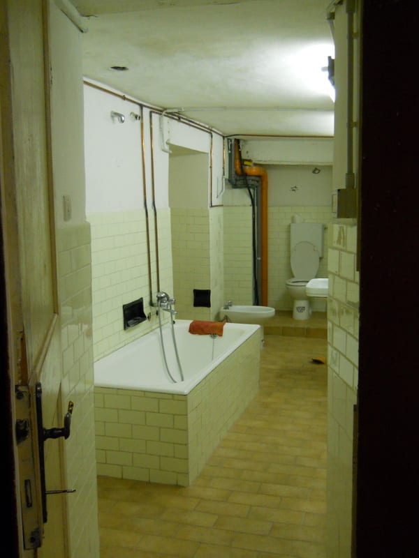 A bathroom before renovation, with white tile, an old bathtub, and exposed copper plumbing visible through a doorway.