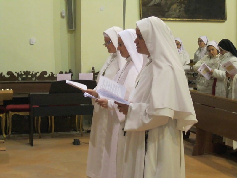 Three sisters in white habits stand in a row in a historic church, holding booklets and singing or reading aloud.